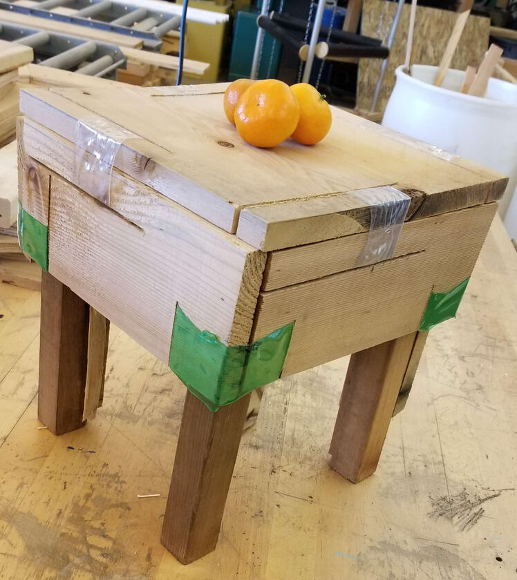 A square wooden stool held together with heat-shrunk plastic bottle scraps around slits in the wood panels. 3 Cuties oranges rest on top for scale.
